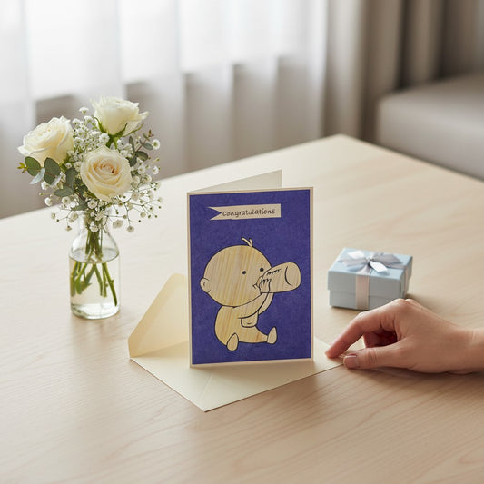 Lokta Cards congratulations card featuring a hand-carved baby drinking from a bottle on a blue textured background.